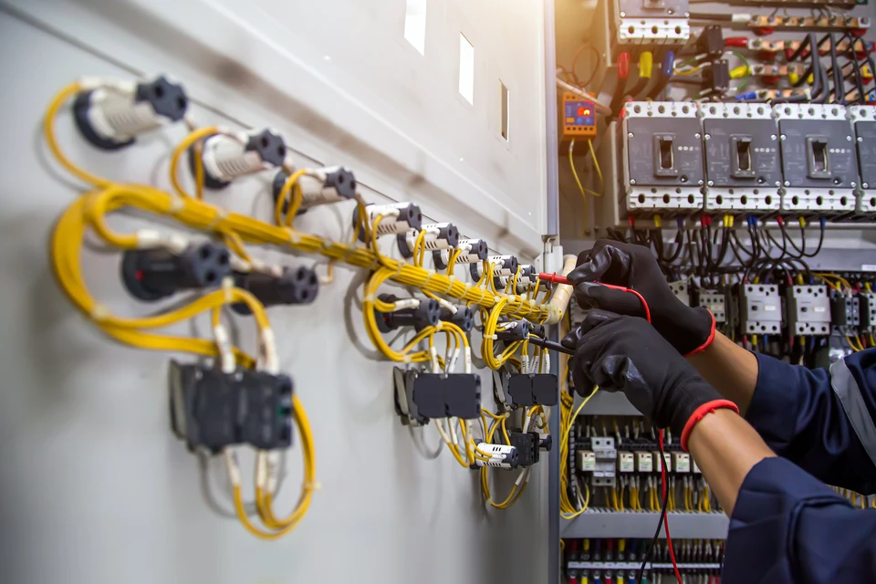Technicien &eacute;lectricien travaillant sur tableau &eacute;lectrique industriel, portant gants de protection noirs avec liser&eacute;s rouges