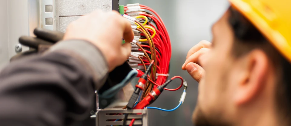Technicien en uniforme travaillant sur installation électrique industrielle, manipulant faisceaux de câbles multicolores (rouge, jaune, bleu, vert) dans tableau de distribution